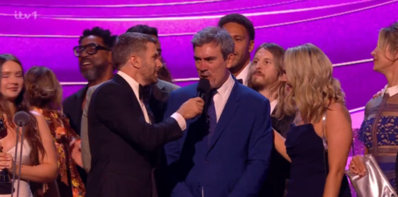 A group of people on stage, two men in the foreground speaking into a microphone.