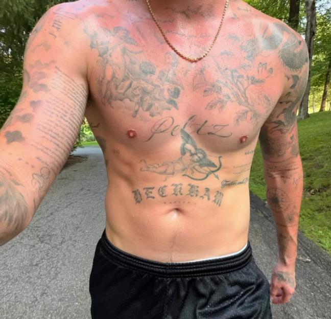 A man with a sunburnt chest and arms covered in tattoos, wearing black shorts and a gold chain, shows off tattoos of "Pelz" and "Beckham" on his chest.