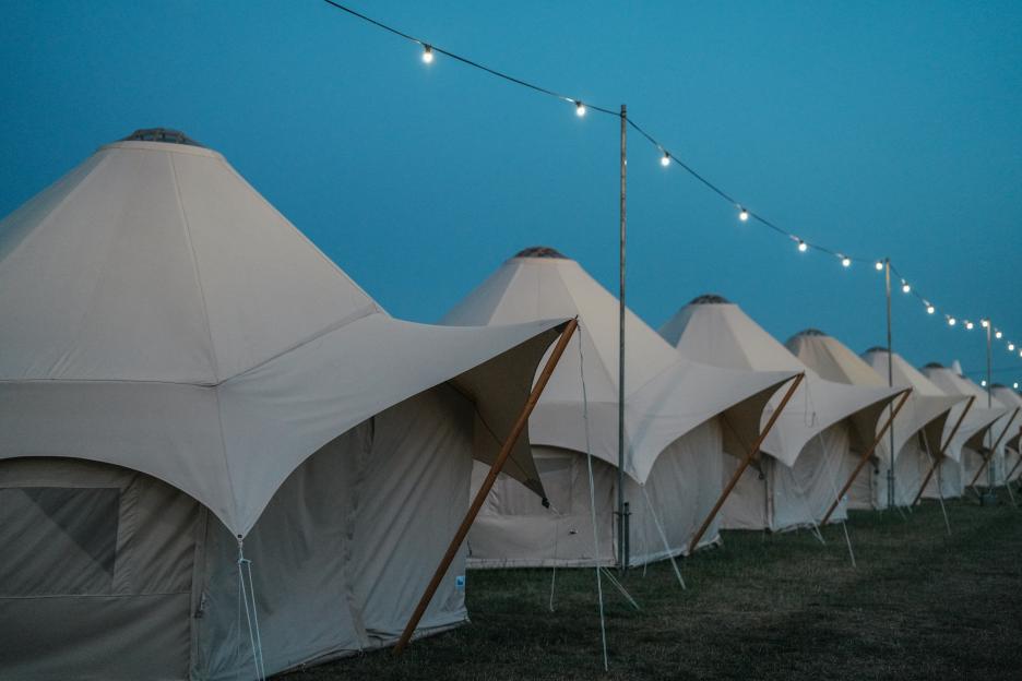 Row of large tents in an open field, with string lights overhead, under a blue twilight sky.