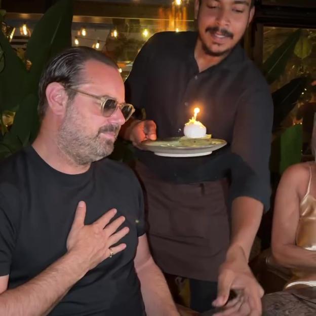 Danny Dyer celebrating his birthday in a restaurant, with a waiter holding a small cake with a lit candle.