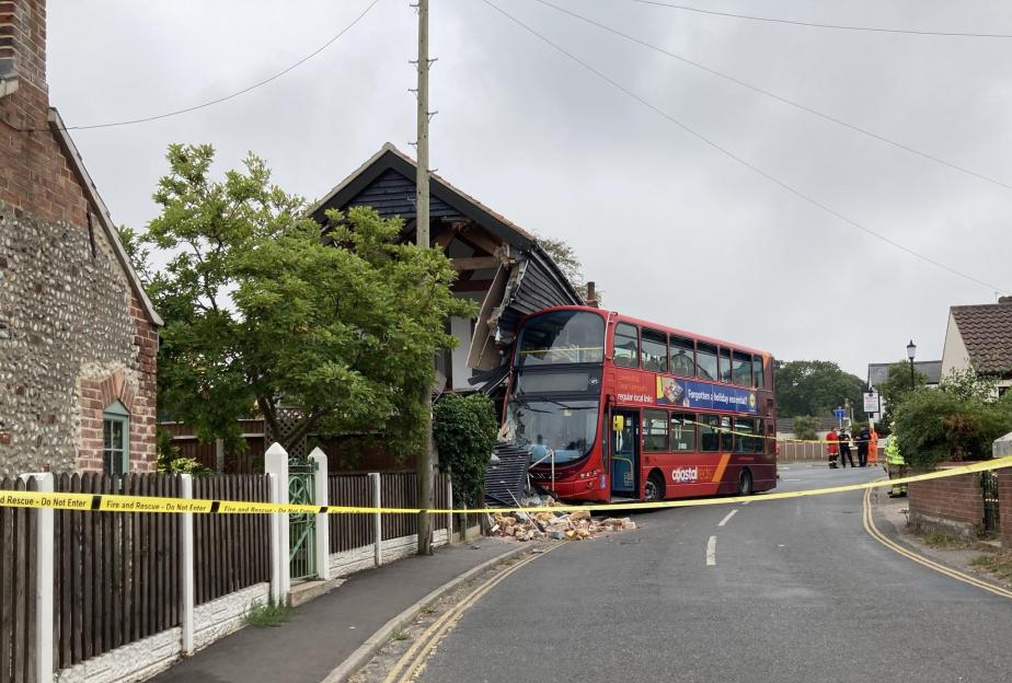 A red double-decker bus crashed into the side of a house, leaving debris on the road, with caution tape across the scene.