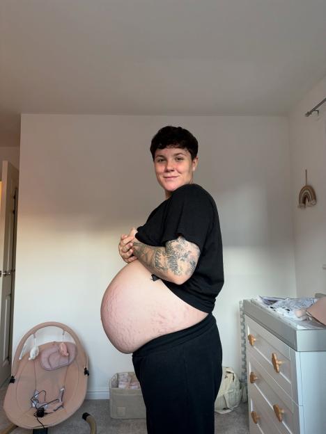 A pregnant person with tattoos on their arms stands sideways, showing their baby bump with stretch marks.