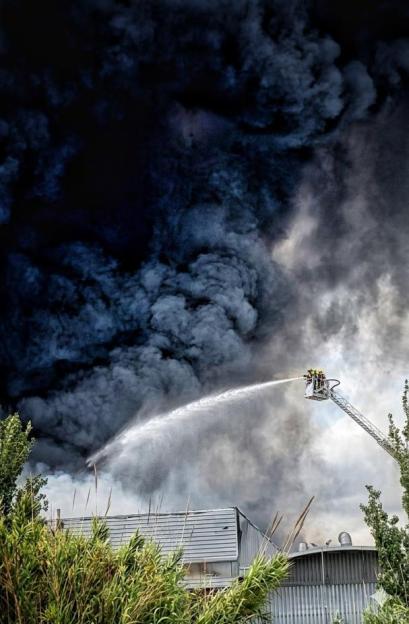 Firefighters in a crane spraying water on a large fire producing thick black smoke above a building.