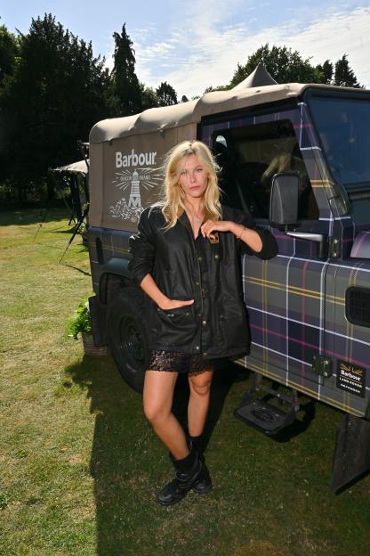 A woman in a Barbour jacket and black boots leans against a tartan Land Rover.