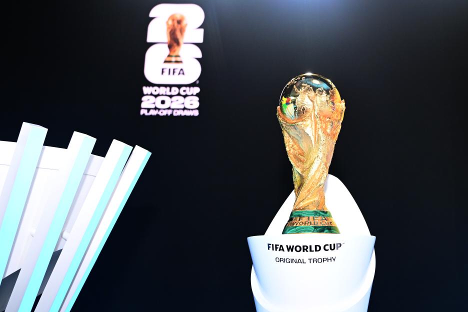 FIFA World Cup trophy on display with "FIFA World Cup 2026 Play-Off Draws" on a screen in the background.