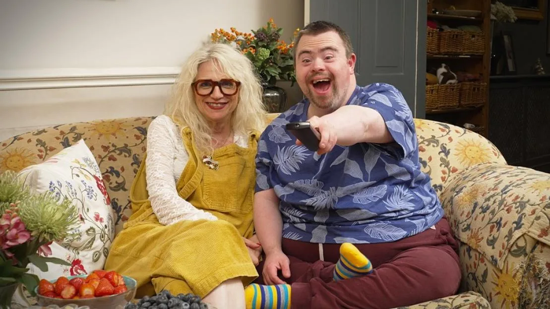 A woman and man with Down syndrome smile on a couch, the man holding a remote.