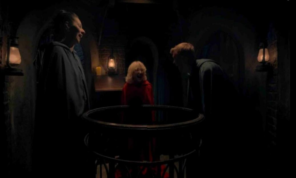 Three contestants from the show "The Traitors" in a dark dungeon-like room with a cauldron-like object in the center.