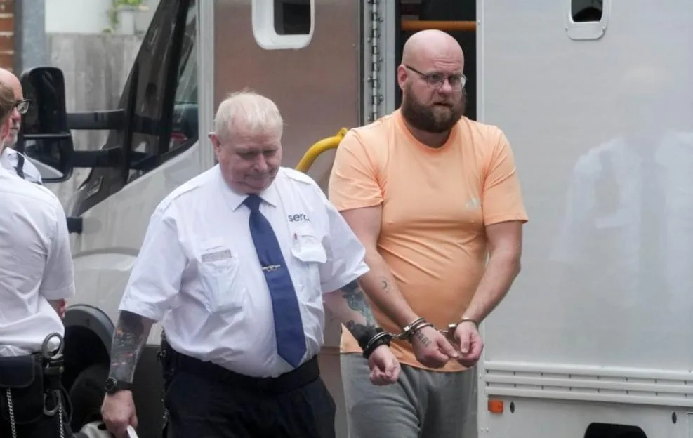 A bald, bearded man in glasses and an orange shirt, handcuffed to a security guard, walks away from a white van.