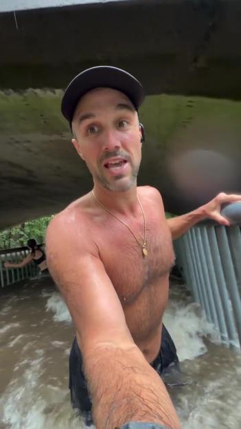 A man wearing a baseball cap stands shirtless in chest-high floodwater, holding onto a railing, with another person further back in the water.