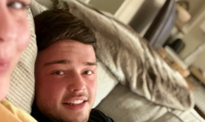 A close-up selfie of a young man smiling with another person's blurred face visible on the left.