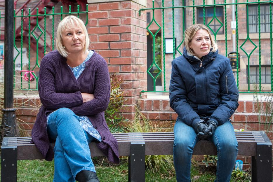 Two women sitting on a bench in a tense conversation, with a green metal fence and brick wall behind them.