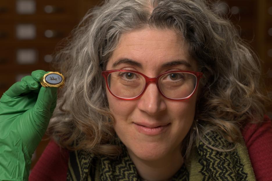 A woman in red glasses and a green glove holds up a gold ring with a black gemstone.