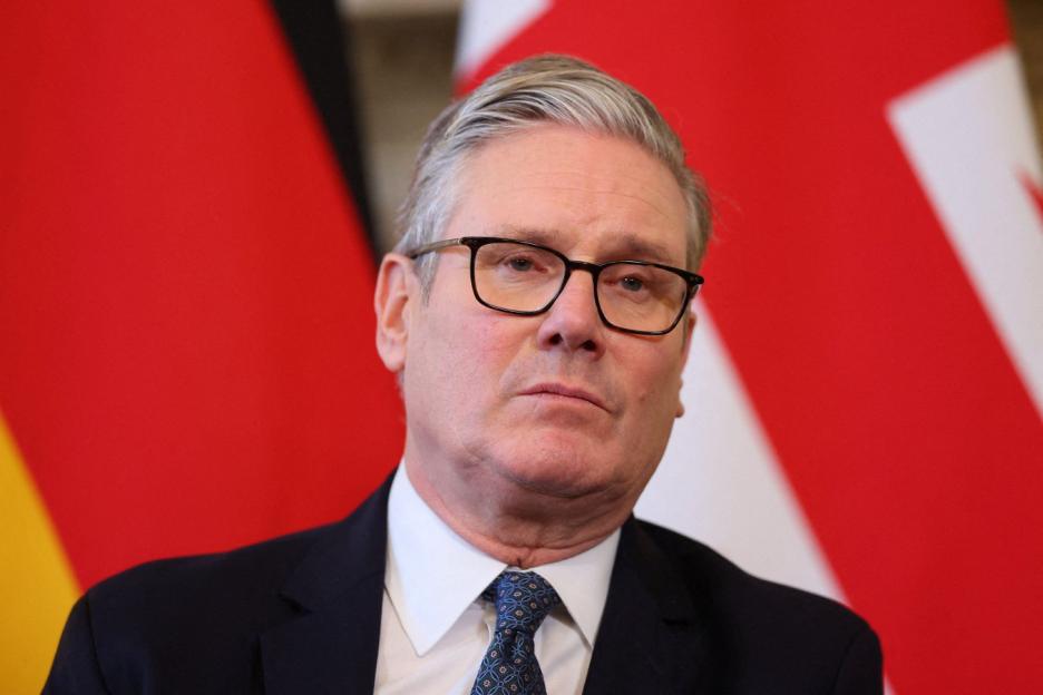 Keir Starmer wearing glasses, a dark suit, and a blue patterned tie, with a red flag partially visible in the background.