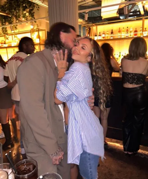 Jesy Nelson posing with her boyfriend, Ben, who is kissing her cheek, in front of a bar.