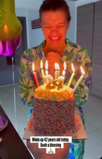 A woman holding a birthday cake with candles that spell "24", though text indicates she is 42.