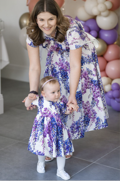 Ilona Andrews helping her baby girl, dressed in matching floral dresses, walk.