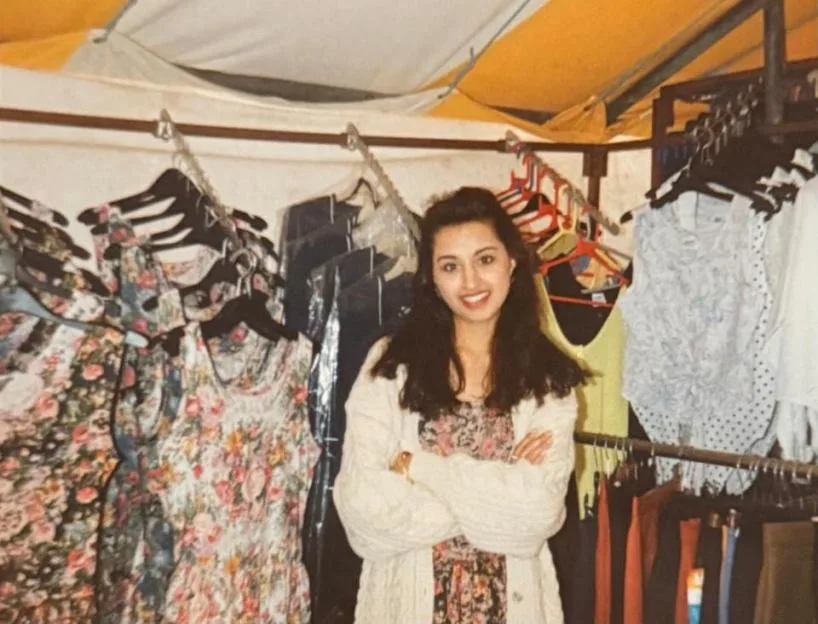 Jinda Mann smiling while standing inside a clothing market stall, surrounded by racks of clothes.