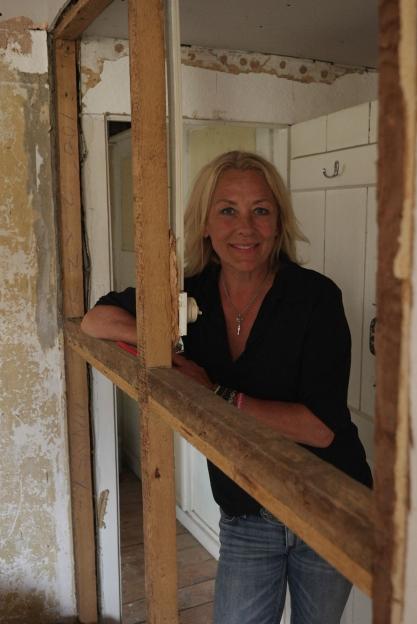 Sarah Beeny smiling while leaning on a wooden frame in a house under renovation.