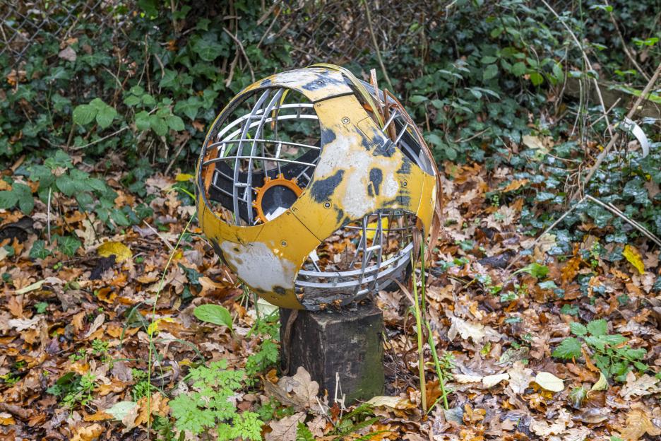 Public art installation resembling a broken globe, made of yellow, black, and white metal, resting on a wooden stump in a garden.