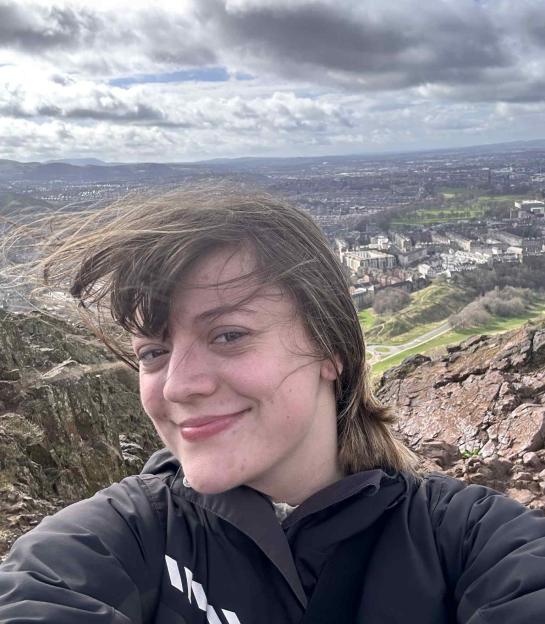A young woman with wind-blown hair smiles at the camera with a city landscape in the background.