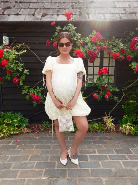 Lauren Layfield in a yellow dress with a pearl bag, standing in front of a building with red climbing roses.