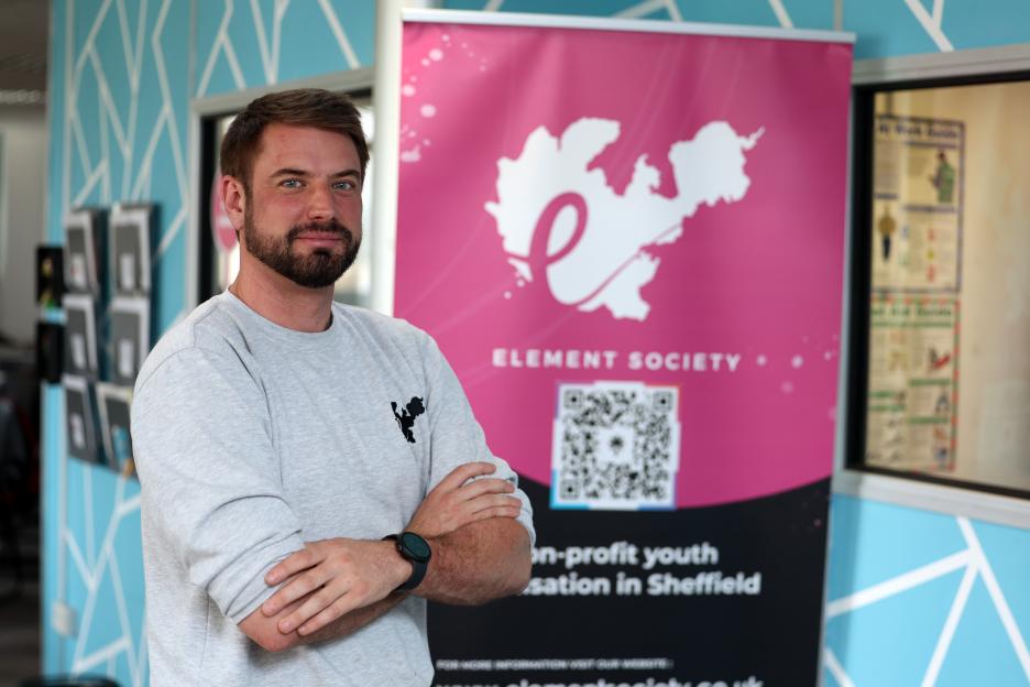 Chris Hill, from Element Society, stands with arms crossed in front of a pink banner for the non-profit organization.