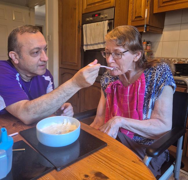 John feeding Gilly, his wife, a spoon of food as she sits in a wheelchair at a wooden table in a kitchen.