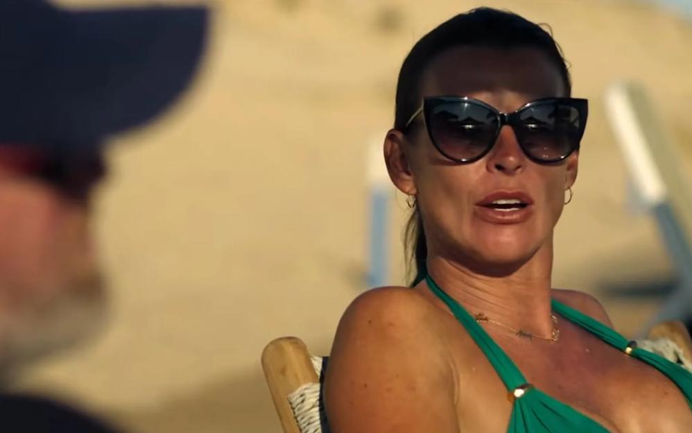 Coleen Rooney in a green bikini and sunglasses, speaking to Wayne Rooney on a beach.