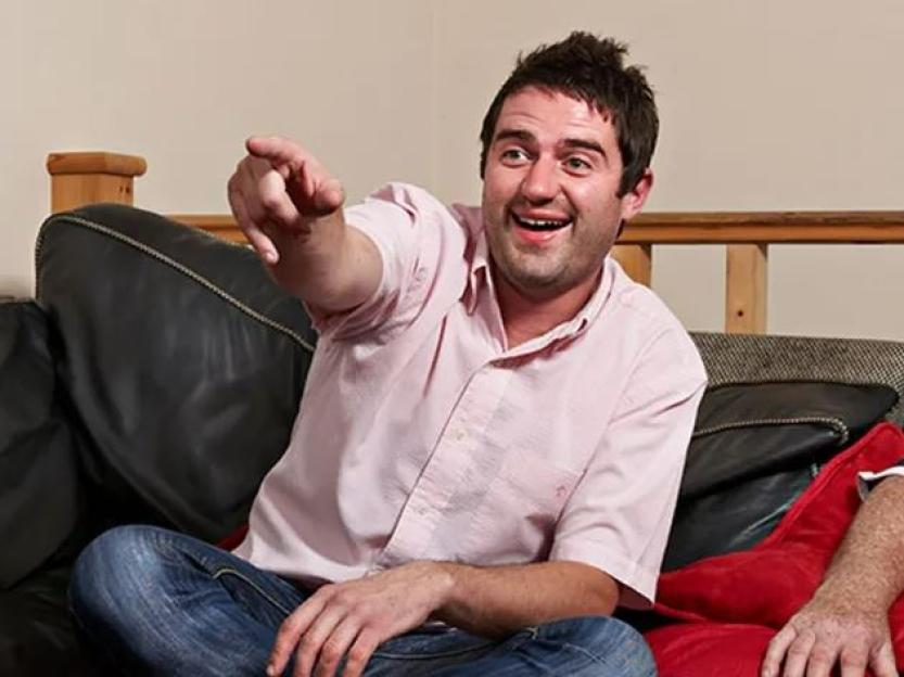 George Gilbey from Gogglebox laughing and pointing while sitting on a couch.