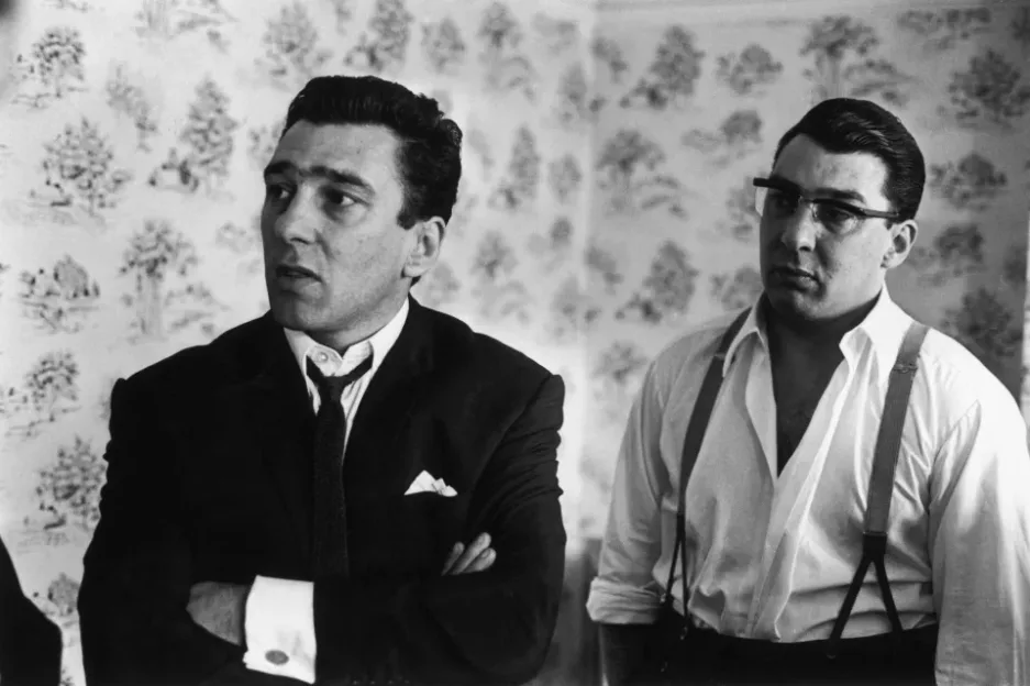 Black-and-white photograph of two men indoors, with patterned wallpaper in the background.