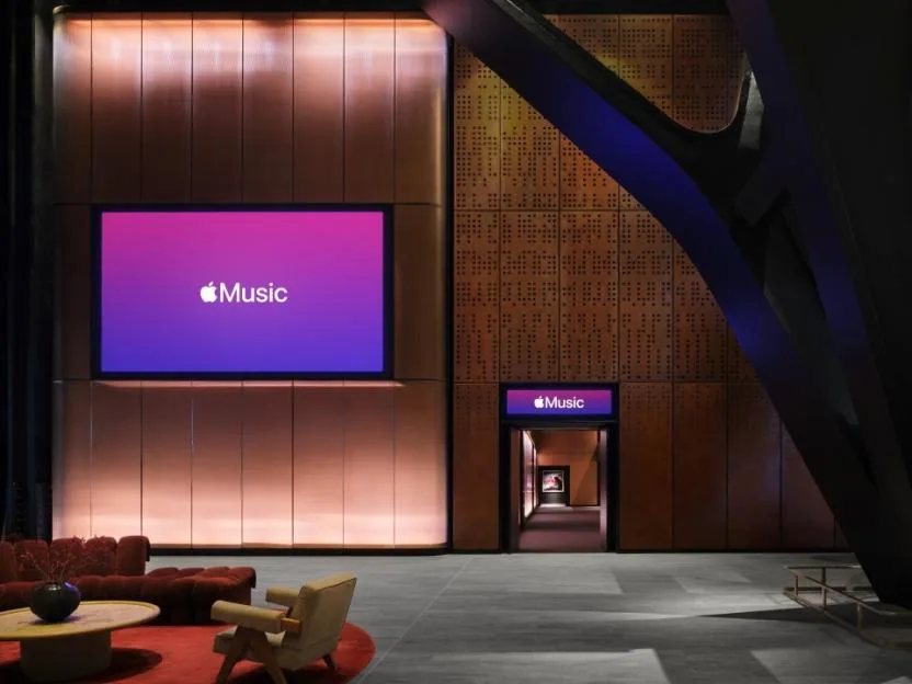 The foyer of Apple Music Hall with a large screen displaying the Apple Music logo, a smaller screen above a doorway leading to a hallway, and red couches with a coffee table on a red rug.