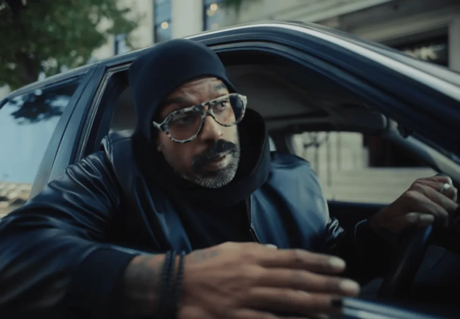 Romesh Ranganathan wearing a black beanie and glasses while sitting in the driver's seat of a car.