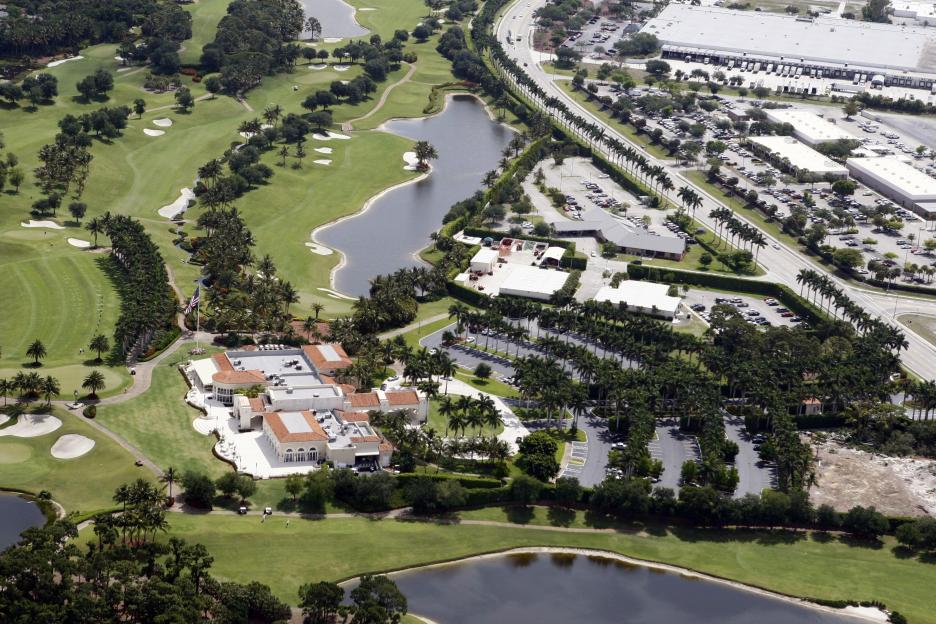 Aerial view of Trump International Golf Club in West Palm Beach, showing the golf course, lakes, clubhouse, and parking lots next to a highway.