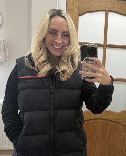 A smiling blonde woman in a black puffer vest takes a mirror selfie with her phone, which holds British pound notes in its clear case.