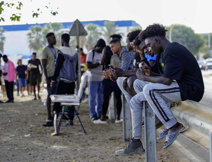 Migrants use cell phones while sitting on a roadside barrier in Ceuta.
