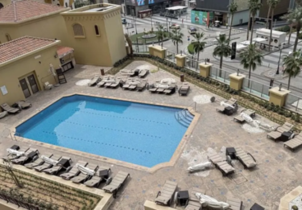 An outdoor swimming pool surrounded by lounge chairs and buildings, with a street and palm trees in the background.