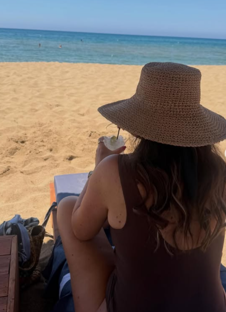 A person wearing a sun hat and swimsuit sits on a beach chair, holding a drink and looking out at the ocean.