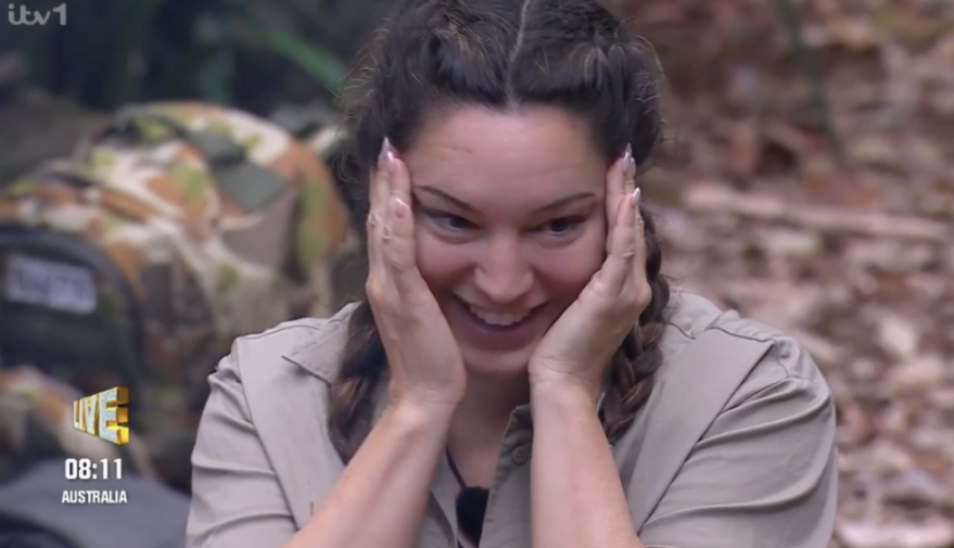 A woman with dark hair in braids smiles with her hands on her cheeks.