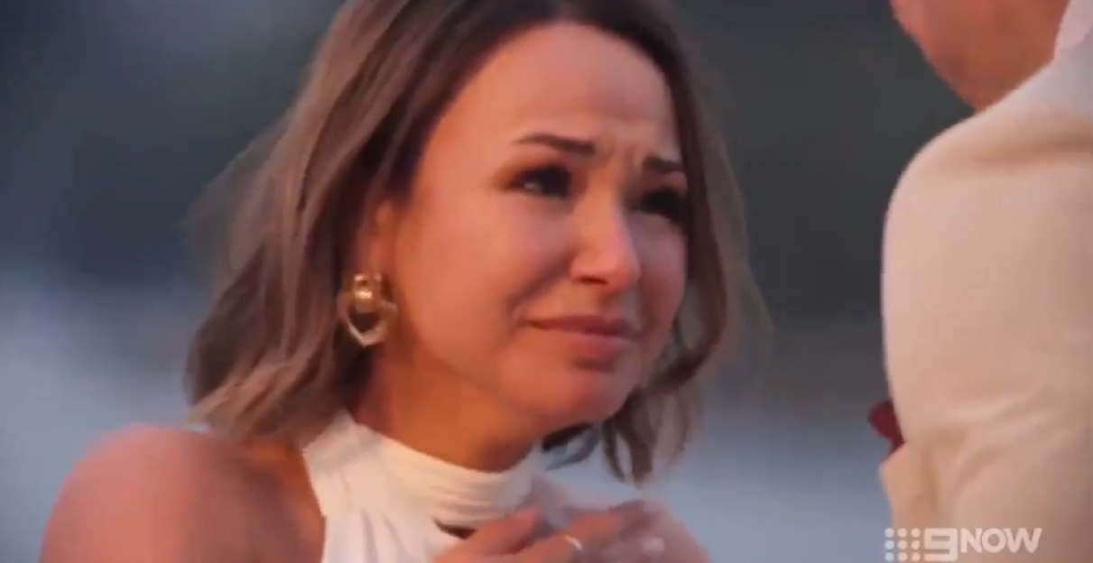 A crying woman in a white dress with her hands to her chest.