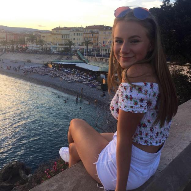 A young woman with long blonde hair and sunglasses on her head smiles at the camera while sitting on a wall overlooking a beach and town.