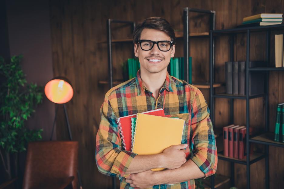 Man in a checkered shirt holding three notebooks while smiling at the camera in a library.