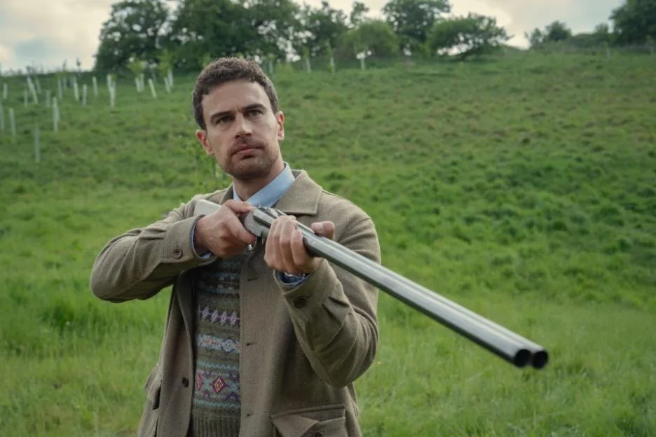 Theo James holding a shotgun in a grassy field.