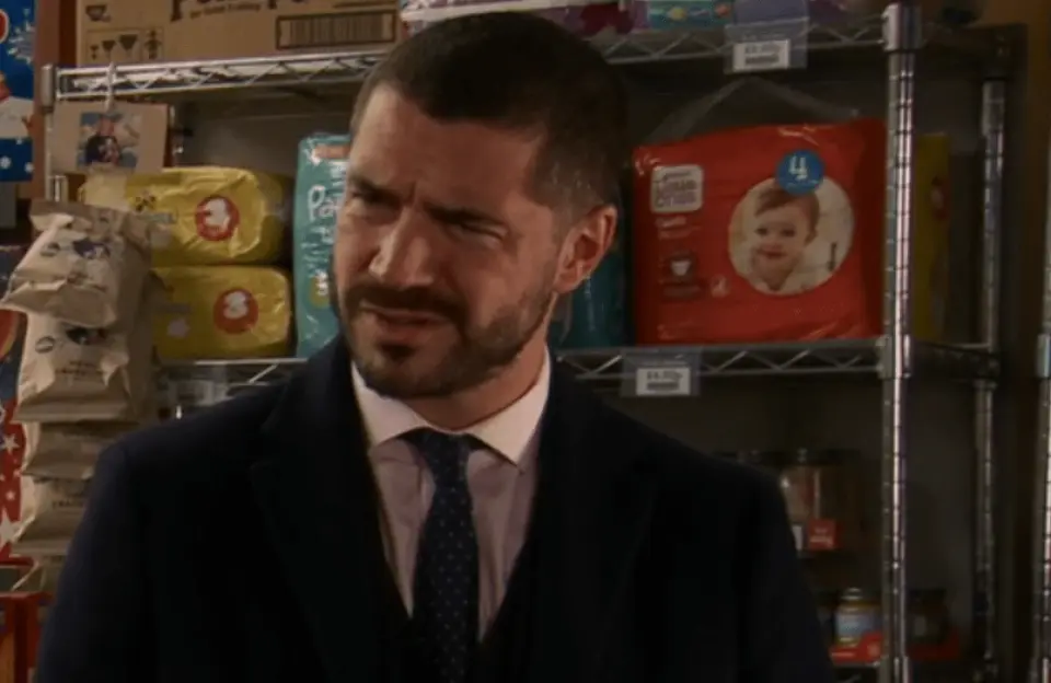 Man with a beard and suit looking displeased in front of shelves with diapers and other items.