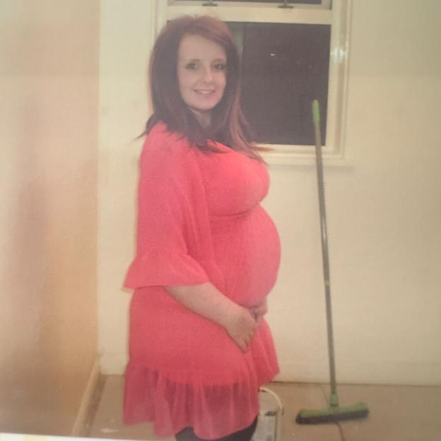 A pregnant woman in a red dress with brown hair smiles, her hand resting on her stomach.
