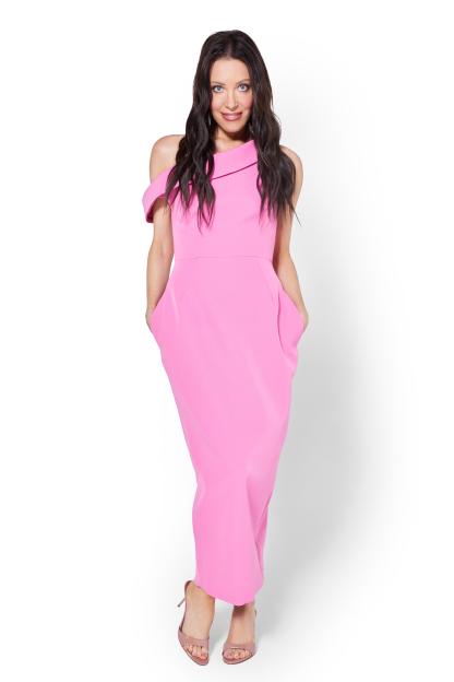 A woman in a long pink dress with an asymmetrical neckline and pockets stands smiling.