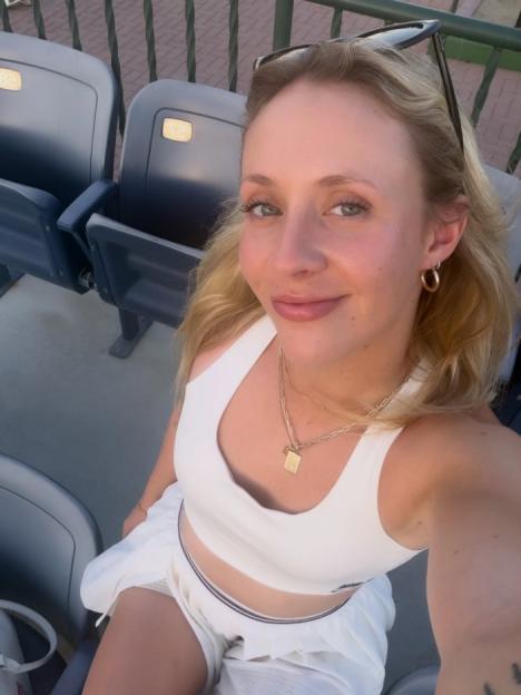 A woman with blonde hair wearing a white top and skirt sits in stadium seating.