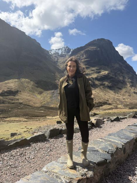 A woman in a green jacket and rain boots stands in front of mountains with snow-capped peaks.