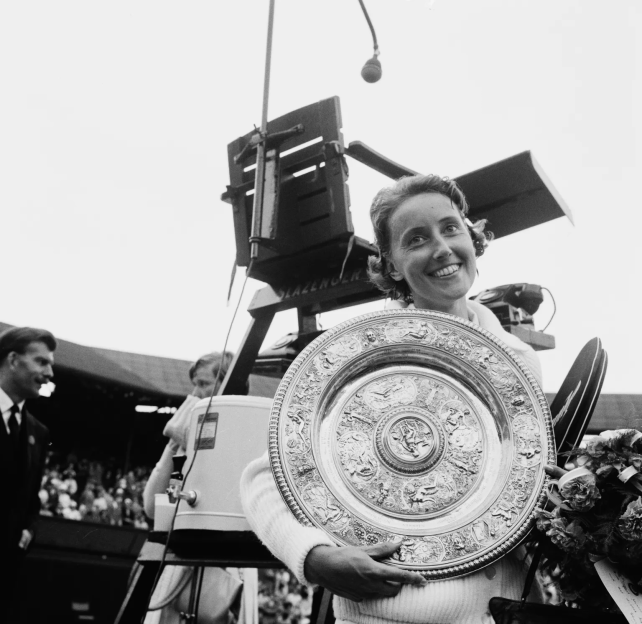 A smiling woman holding a large, ornate silver Wimbledon trophy and a bouquet of flowers.