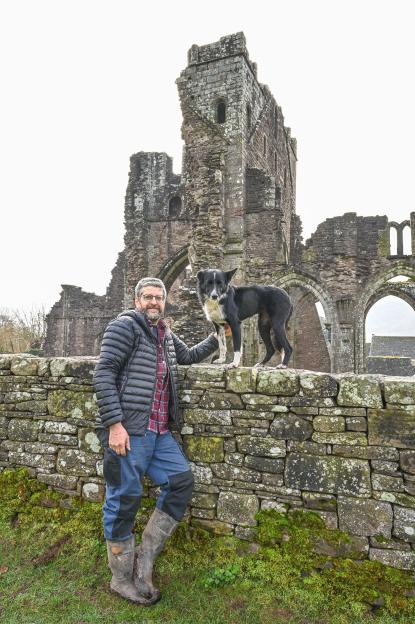 Farmer Mark Morgan stands with his dog on a stone wall in front of the Llanthony Priory ruins, worried about the lambing season due to road closures.
