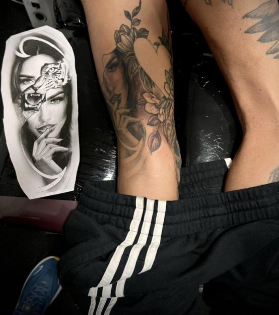 A person's leg featuring a tattoo of a woman's face intertwined with a floral design and a heart, alongside a printed tattoo design of a woman's face merged with a tiger's face.